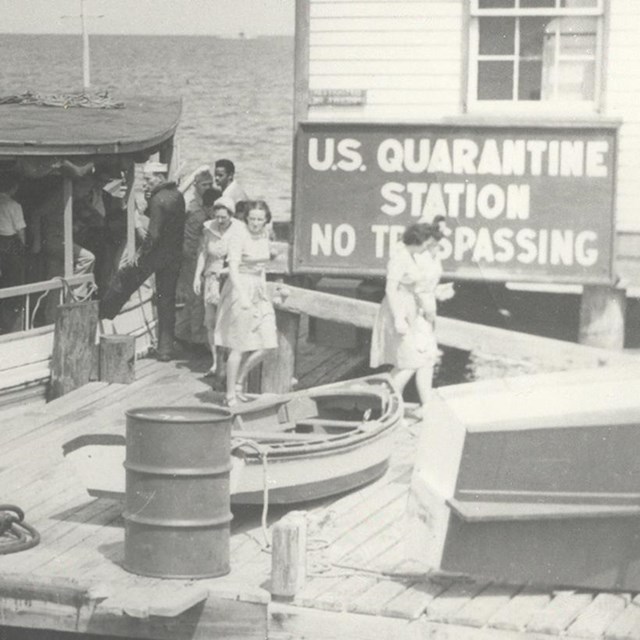 Black and white image of a boat docked at a small wooden pier and building.