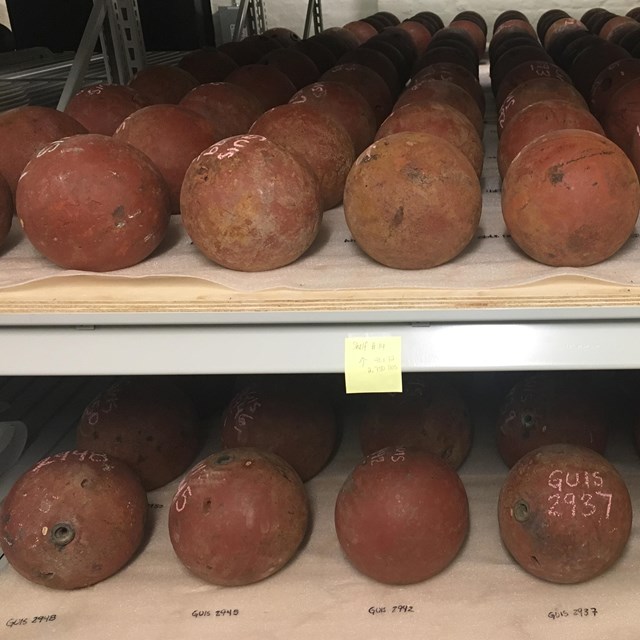 Two metal shelves are filled with historic cannonballs neatly arranged.