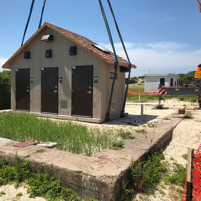 A crane lowers a restroom structure on to a foundation in a park setting.