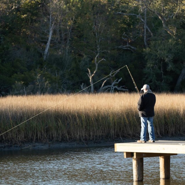 A fisherman casts a line into the bayou from a pier.