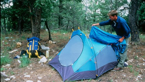 A man sets up a blue tent at a wilderness campsite