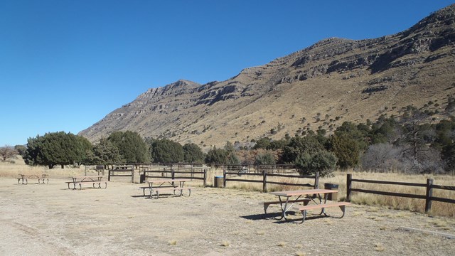 Picnic tables, RV sites, and horse corrals are located below a high desert mountain cliff. 