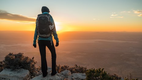 A hiker stands on a mountain top at sunset