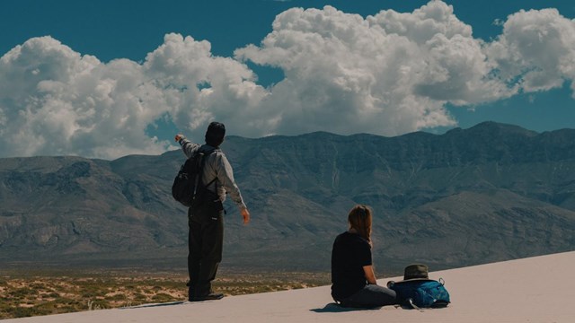 A park ranger and visitor on a white gypsum sand dune with tall mountains in the background. 