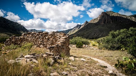 A trail leads past a ruined stone wall with desert mountains behind.
