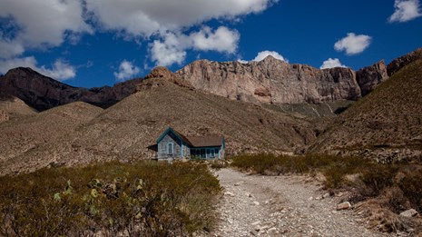 A dirt road leads to a small ranch house dwarfed by desert mountains