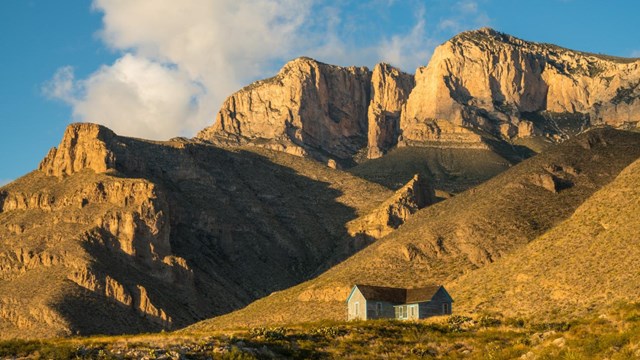 A small blue ranch house sits below tall yellow mountain cliffs. 