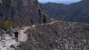 A backpacker walks along a stony trail in a steep desert mountain landscape