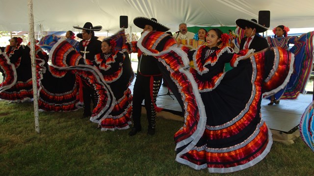 A group of men and women performing traditional dances with a band in the background. 