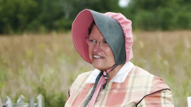 A woman wearing a pink plaid historic period dress with pink, green bonnet.. 