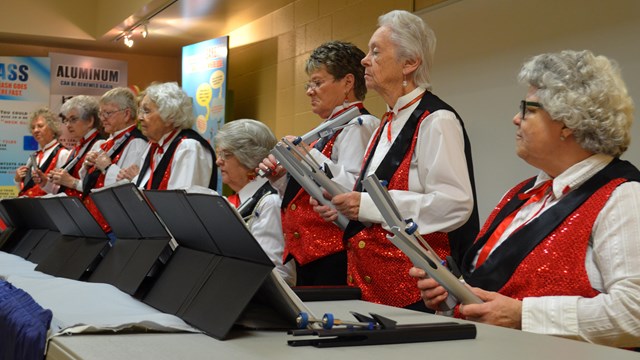 8 women wearing red and black vests, white shirts, and red bow ties playing handbells.