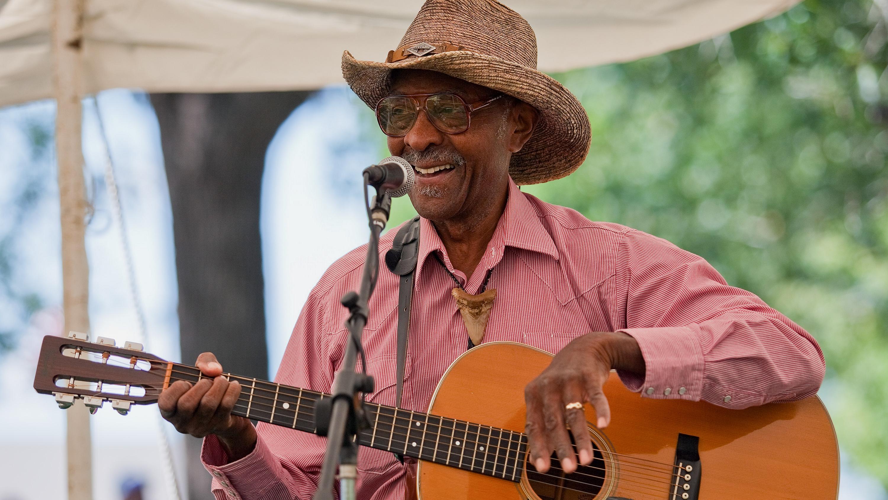 A man playing a guitar.  He is wearing red shirt, blue jean, and a tan cowboy hat.