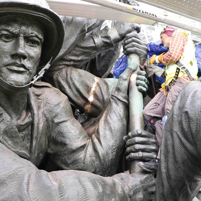 A worker suspended from scaffolding works on a bronze statue of a marine.