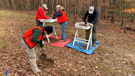 A group of archeologists screening soil from a nearby shovel hole against a winter forest backdrop.
