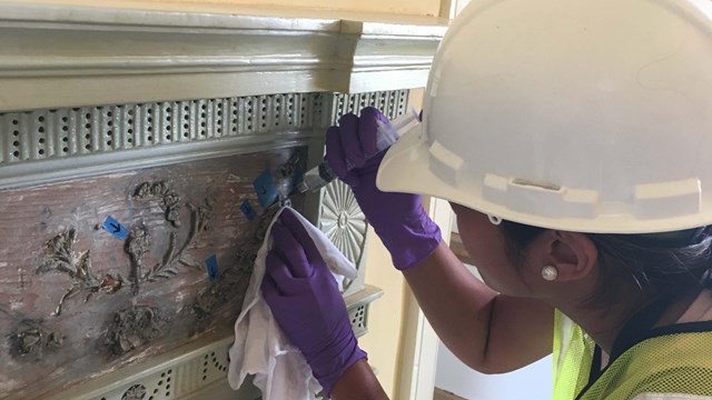 A woman in a hardhat makes repairs to an ornate wooden mantelpiece