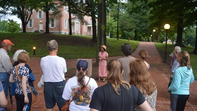 A young African American woman in 1930s garb stands near a monument; visitors listen to her talk