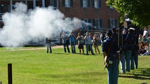 men depicting Civil War soldiers fire blackpowder small arms weapons; people watch from behind ropes