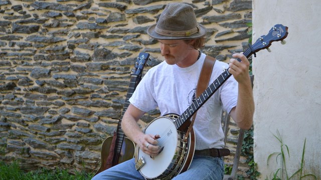 man playing the banjo; sitting in front of a stone wall