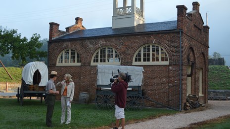 ranger interviewed by newswoman; man focuses a TV camera on them; backdrop is John Brown's Fort