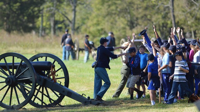 kids with hands raised next to a ranger, a volunteer dressed as a Civil War soldier, and a cannon