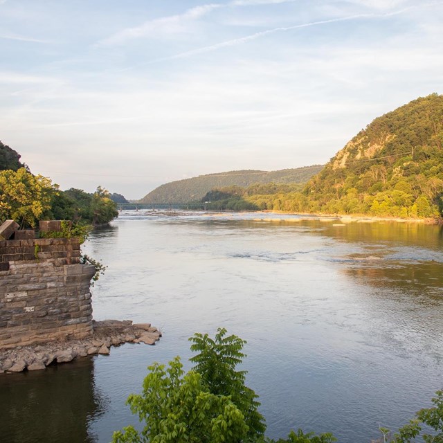 Wide river in between wooded hills, with ruins of bridge supports emerging from the water