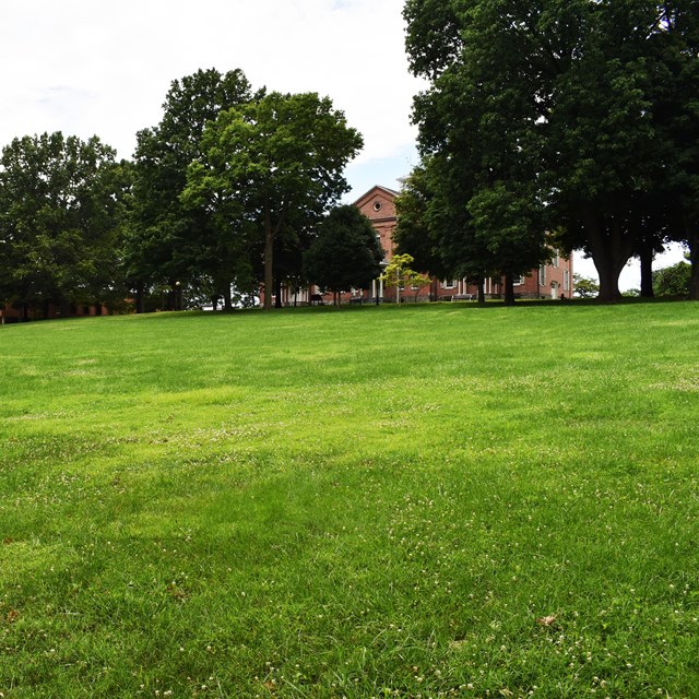Large grassy area in front of two-story rectangular brick building surrounded by trees