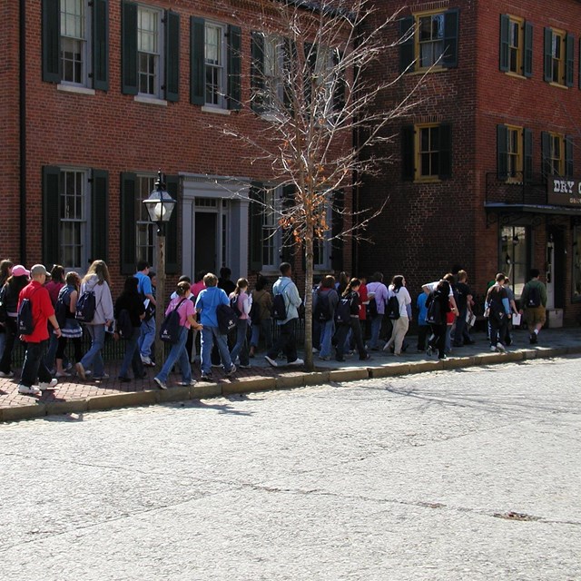 Group of school-age youth walk on town sidewalk past brick buildings