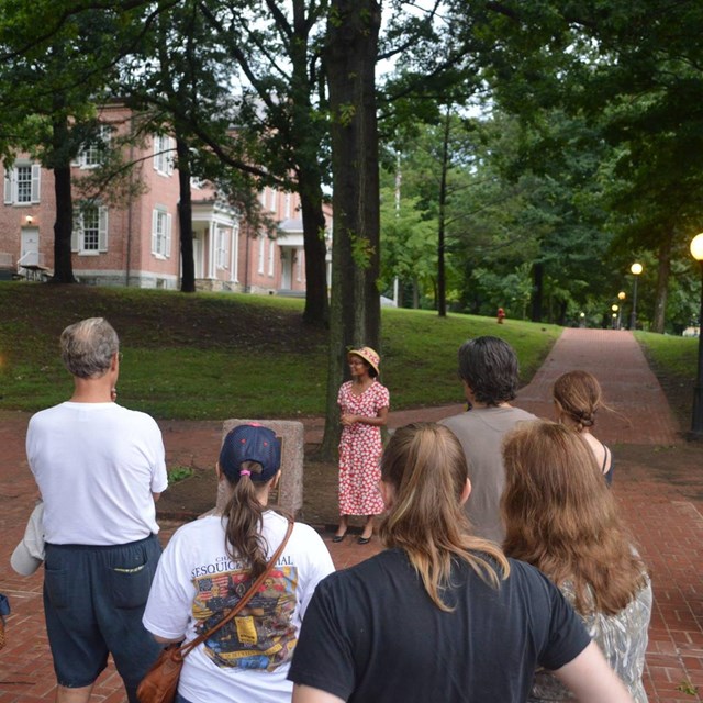 A young African American woman in 1930s garb stands near a monument; visitors listen to her talk