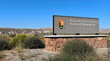 Photo of the Hagerman Fossil Beds entrance sign, with the fossil beds in the distant background.