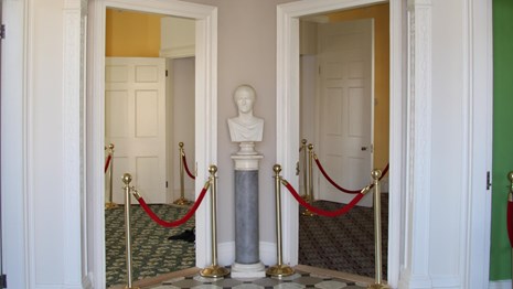 A marble bust of Hamilton stands on a pedestal in a hallway with an arch and checkered flooring.