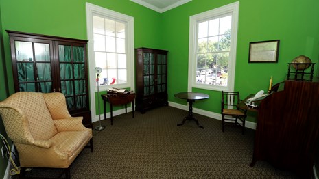 A room painted lime green contains mahogany bookshelves, a winged armchair, a tea table and a desk.