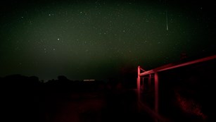 A railing is illuminated by red light. In the background, a shooting star streaks across a dark sky.