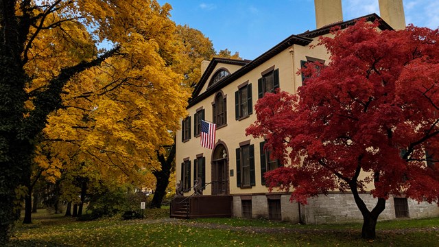 A two-story yellow house with brown trim, surrounded by fall trees.