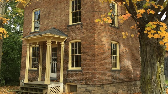 The front and side of a brick two-story house with small white columns in front and fall leaves.