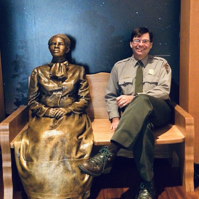 A park ranger sits beside a bronze statue of Harriet Tubman with a Tubman quote above them.