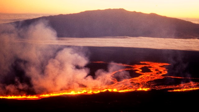 Molten lava flowing down a mountain side with a silhouetted mountain beyond at sunrise