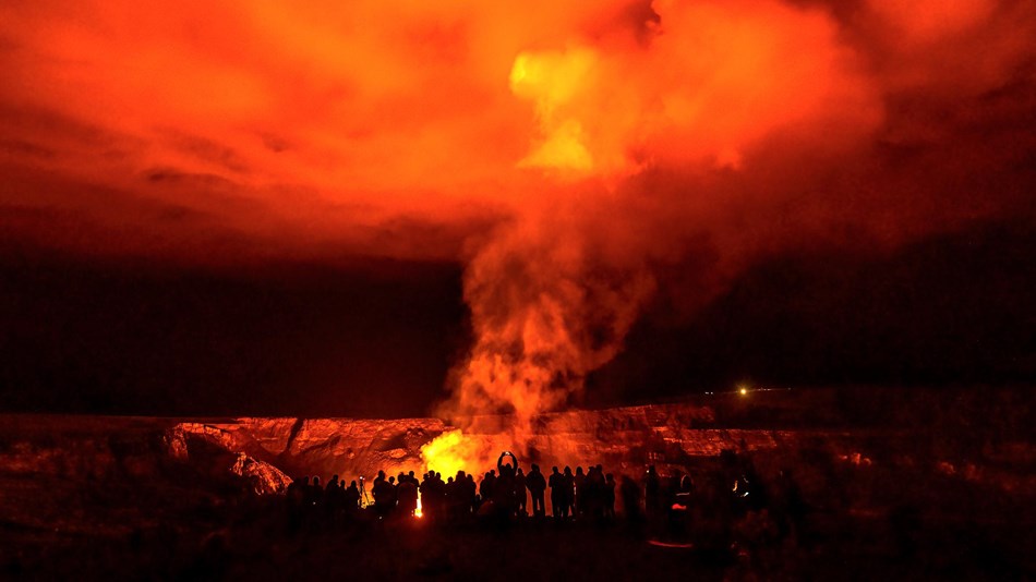 Visitors standing in front of a deep dark crater at night with bright orange glow and smoke.