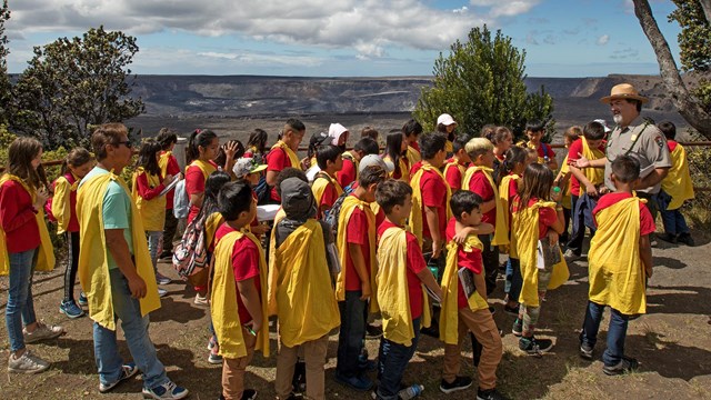 A group of students wearing red and yellow talking to a park ranger in front of a volcanic caldera
