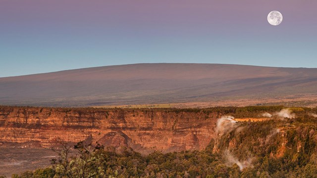 A round mountain below a full moon with a volcanic caldera in the foreground