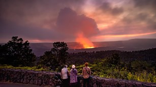Park visitors watching a volcanic eruption from an overlook with a stone wall