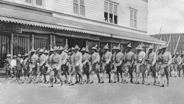 Black and white photo of a company of soldiers marching through a town street