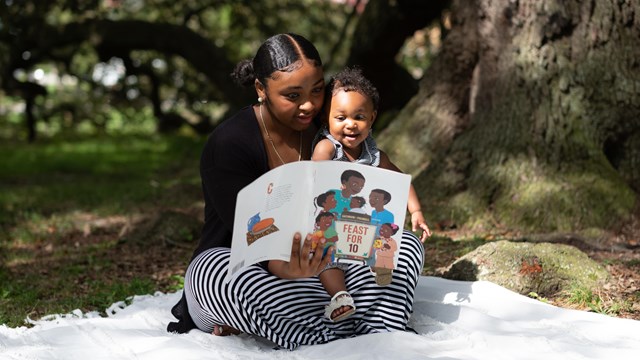 mother reads to baby while sitting on a blanket under a tree