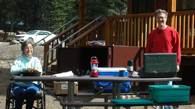 Disabled woman and man standing at picnic table with camping stove, coolers and equipment