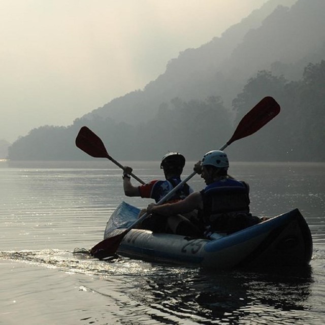 Two people wearing helmets and life jackets paddling in lake on inflatable kayak