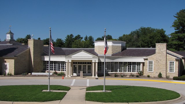 A sprawling one story public building of rough-faced yellowish stone has a white portico entrance.