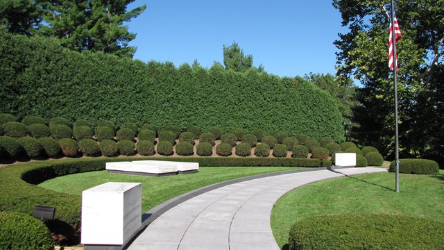 Two plain marble ledgers mark graves in a semicircular planting of shrubs with a US flag.