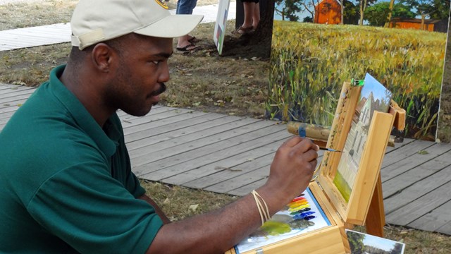 A man seated at an easel paints and displays finished paintings.