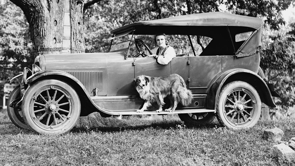 Black and white picture of woman in driver's seat of 1920s car with a dog. Library of Congress photo