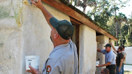 Rangers hold buckets of paint while they reach up to brush paint on a stucco wall.