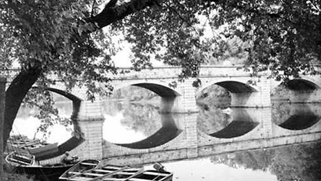 Black and white photo of a bridge over still water.